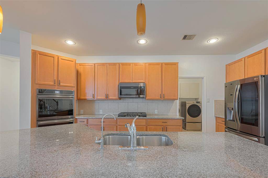 This kitchen features a spacious granite countertop island with a sink, stainless steel appliances, ample wooden cabinetry, and modern lighting. A view of the laundry area is visible through an adjacent doorway, enhancing functionality.