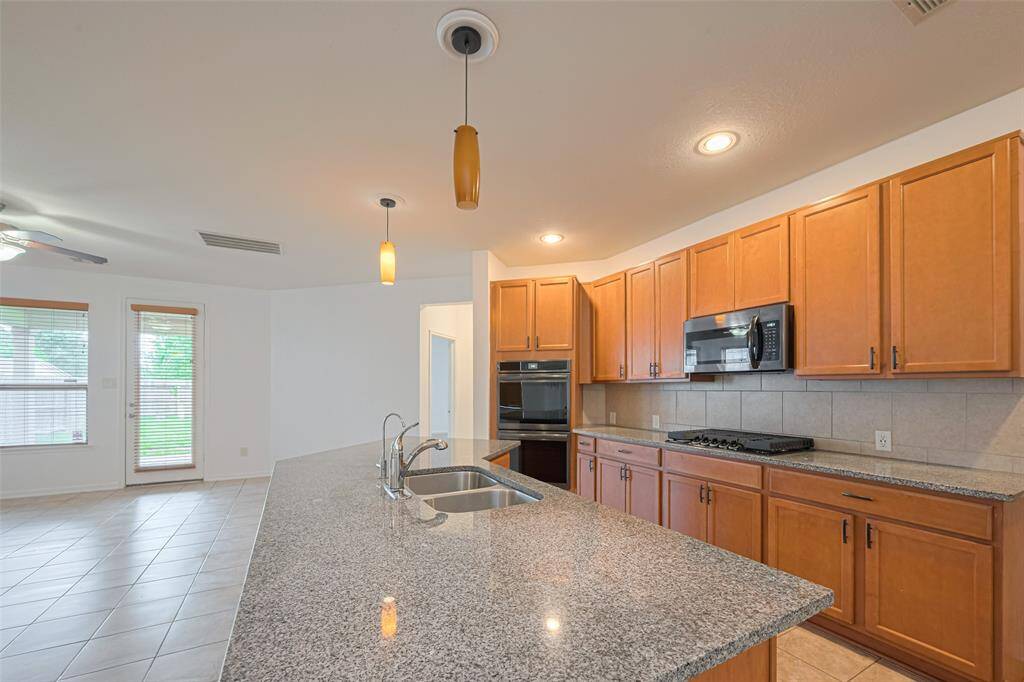 This kitchen features granite countertops, ample wooden cabinetry, and modern appliances, including a built-in oven and microwave. The space is open and bright with tile flooring and a view into the adjacent living area, highlighted by large windows and a ceiling fan.