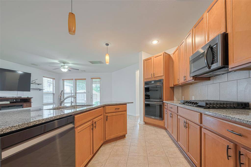 This modern kitchen features granite countertops, ample wood cabinetry, and stainless steel appliances, including a built-in oven and microwave. The open layout connects to a bright living area with large windows and a ceiling fan, creating a spacious and inviting atmosphere.