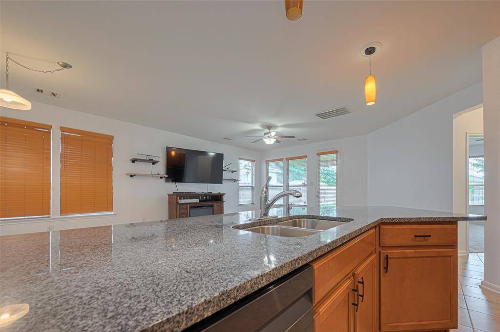 This open-concept living area features a modern kitchen with a large granite countertop island and double sink, transitioning into a bright space with large windows, wooden blinds, and a mounted TV above a fireplace.