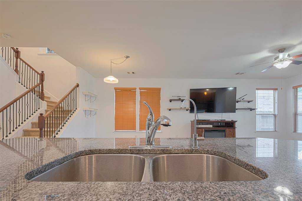This open-concept living area features a modern kitchen island with a double sink and granite countertop, overlooking a cozy living room with large windows, a mounted TV, and a ceiling fan. A stylish staircase with wrought iron railings adds an elegant touch.