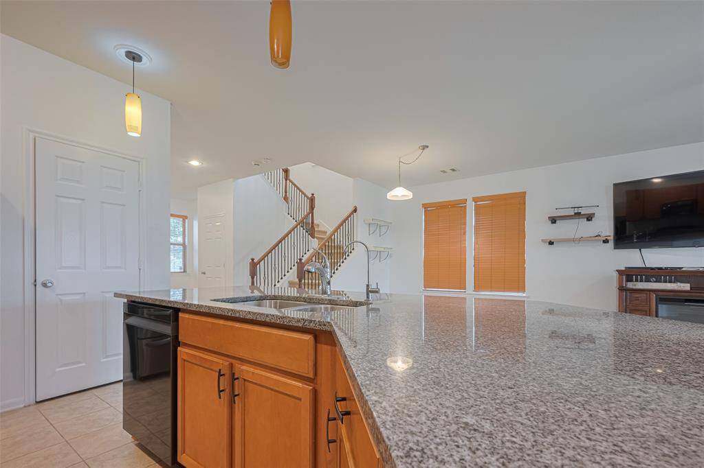 This photo showcases a modern kitchen with a spacious granite countertop island, pendant lighting, and wooden cabinetry. The open layout leads to a living area with large windows, wooden blinds, and a staircase.