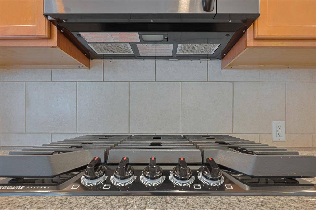 This photo showcases a modern kitchen with a sleek gas cooktop, featuring multiple burners and precise control knobs. It is complemented by a stainless steel range hood and surrounded by light wood cabinetry and neutral tile backsplash for a contemporary look.