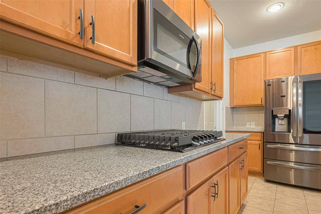 This kitchen features modern stainless steel appliances, granite countertops, and light wood cabinets. The space is accented by a tiled backsplash and floor, offering a sleek and functional cooking area.