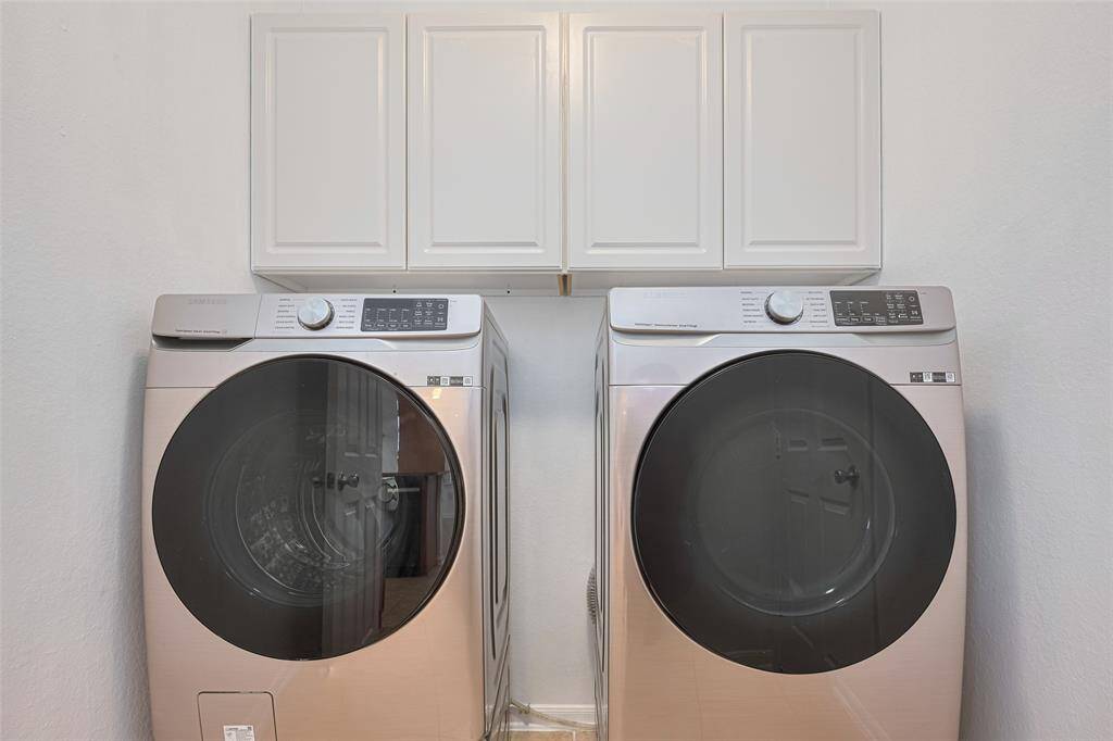 This photo features a modern laundry room with sleek front-loading washer and dryer units. Above them, there are white cabinets providing additional storage space. The room has a clean, minimalist design.