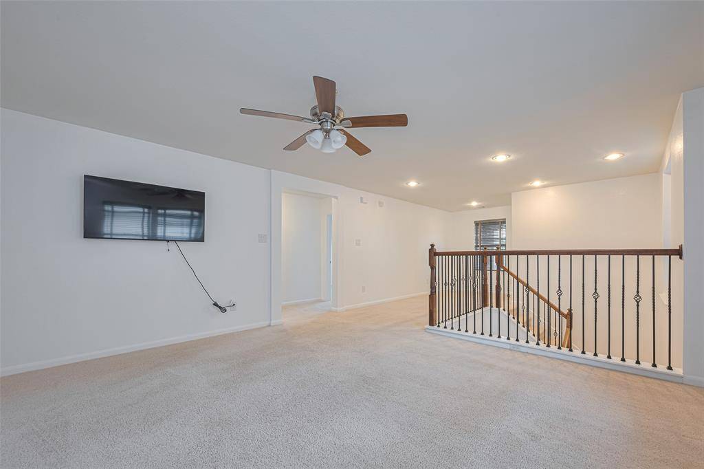 This photo shows a spacious, carpeted upper-level landing area featuring a ceiling fan, mounted TV, and recessed lighting. There’s a railing overlooking the stairs, leading to a bright hallway.