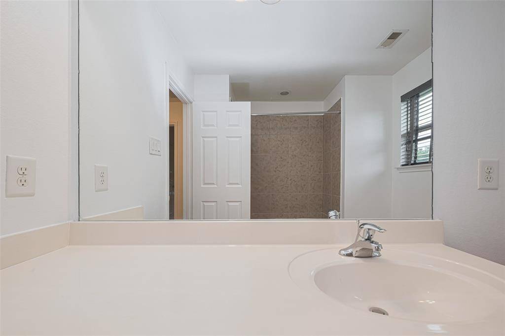 This photo features a clean, modern bathroom with a large mirror above a sleek countertop. There's a single sink with a chrome faucet, ample counter space, and an adjacent shower with tiled walls. Natural light enters through a window with blinds.
