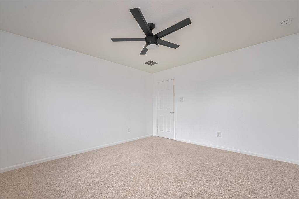 This is a spacious, carpeted room with white walls, featuring a modern black ceiling fan and a single door, ideal for a bedroom or office.