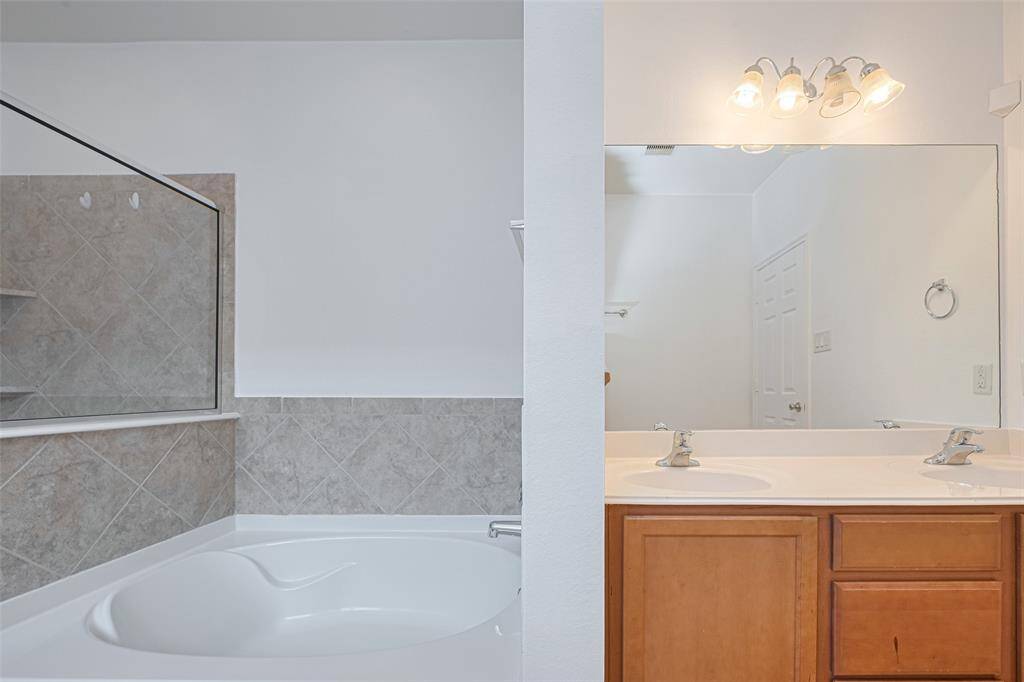 This bathroom features a spacious tub with tiled walls, a large mirror, dual sinks with a wooden vanity, and bright overhead lighting.