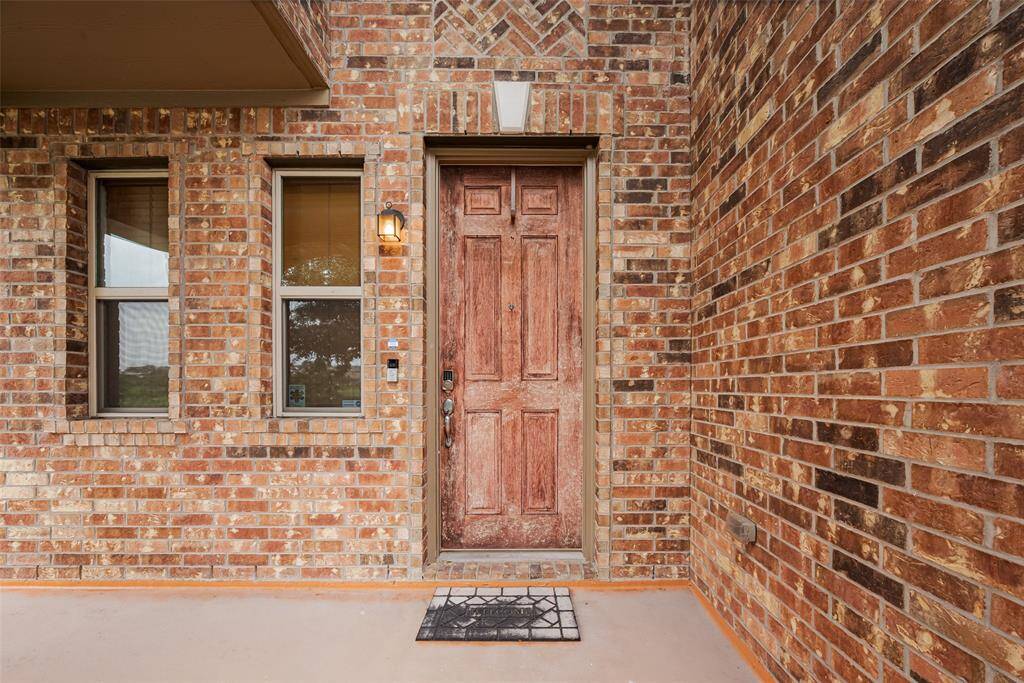 Charming brick entryway featuring a rustic wooden door, flanked by two windows and a warm porch light, creating an inviting and classic look.