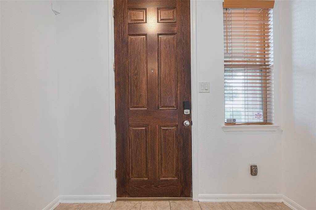 This photo showcases a welcoming entryway with a rich wooden front door, complemented by a window with wooden blinds, set against a backdrop of neutral walls and tile flooring.