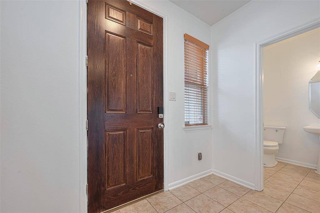 This photo shows a welcoming entryway with a rich wooden front door, tiled floor, and a window with blinds. There's also a view into a small bathroom with a toilet and sink, featuring bright lighting.