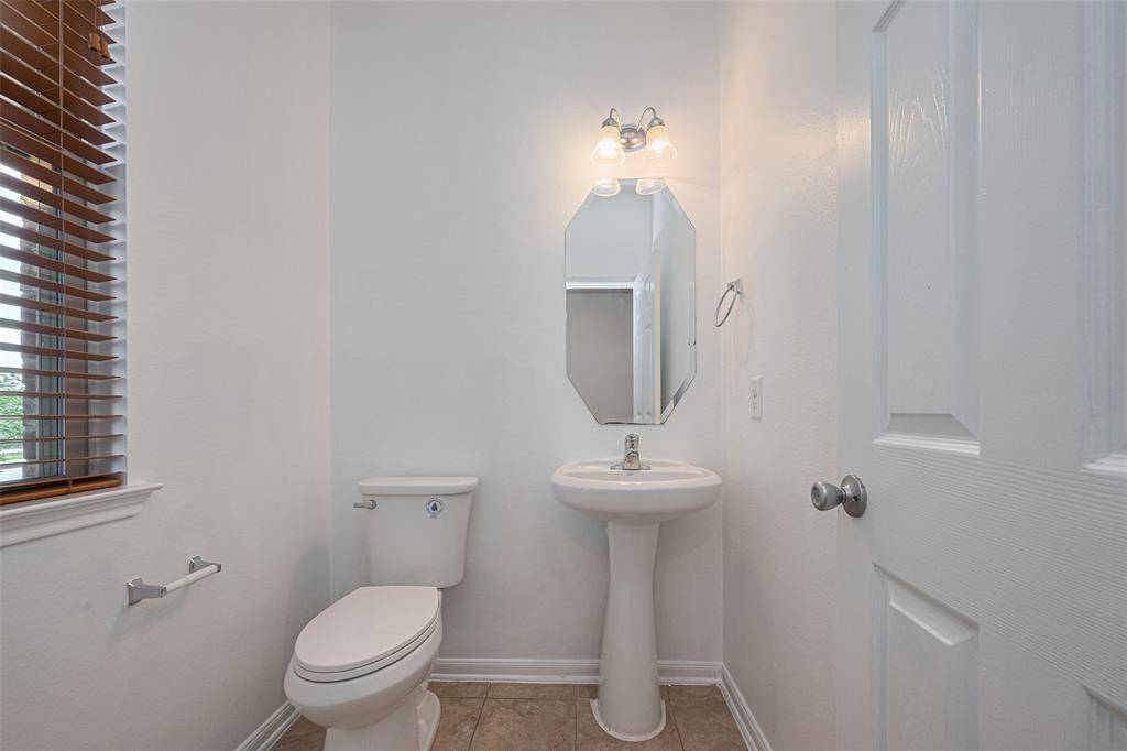 This photo shows a small, bright bathroom featuring a pedestal sink, toilet, and an octagonal mirror. The room has white walls, a window with wooden blinds, and a light fixture above the mirror.