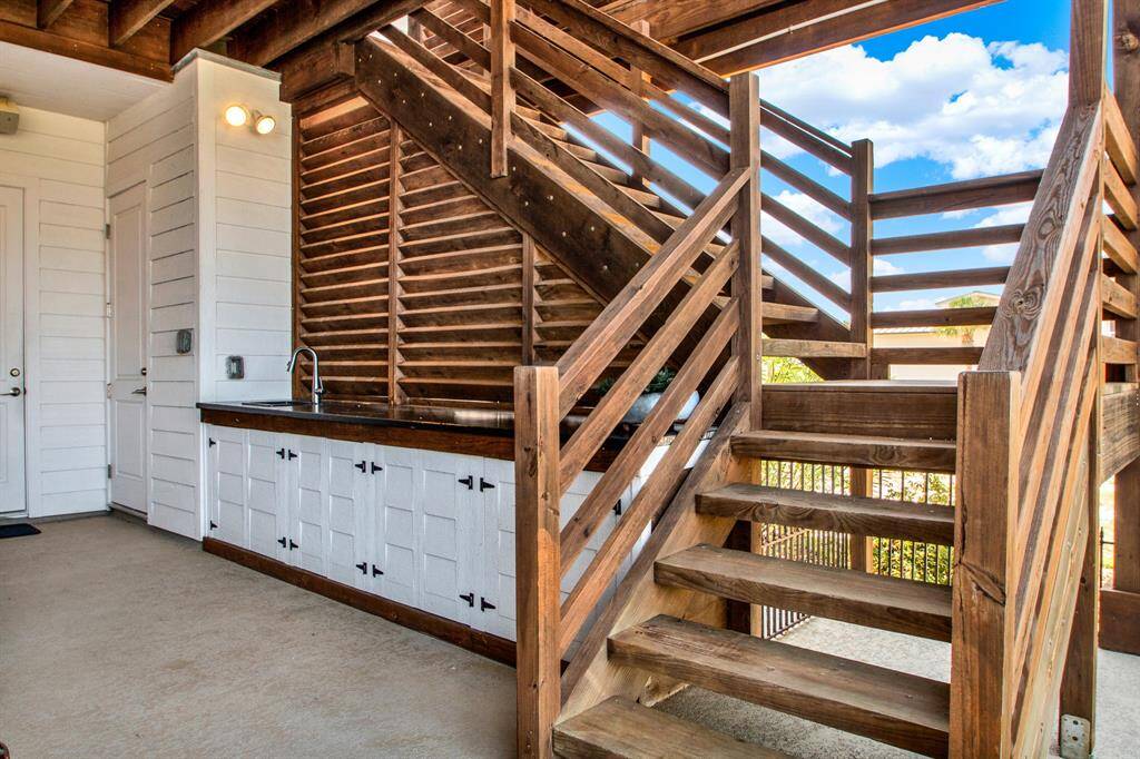 This interior space features rustic wooden stairs leading to an upper level, with a cozy kitchenette underneath, white cabinetry, and a farmhouse sink, set against a backdrop of ample natural light and a view of the outdoors.