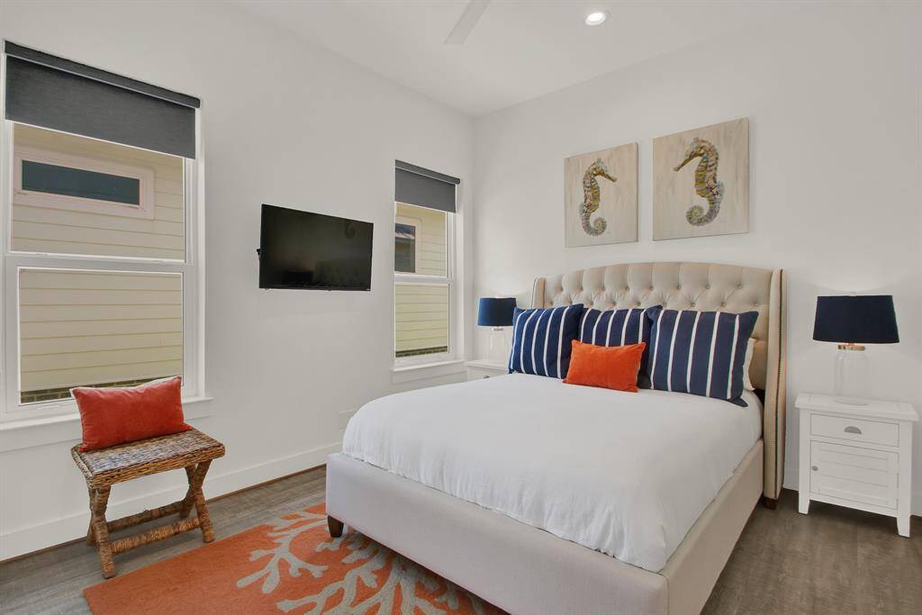 This is a modern, well-lit bedroom featuring a large bed with a tufted headboard, crisp bedding with accent pillows, two windows with dark shades, a mounted flat-screen TV, and coastal-inspired art above the bed. A small, rattan stool and a nightstand with a lamp complete the space, set against neutral walls and a patterned orange rug.