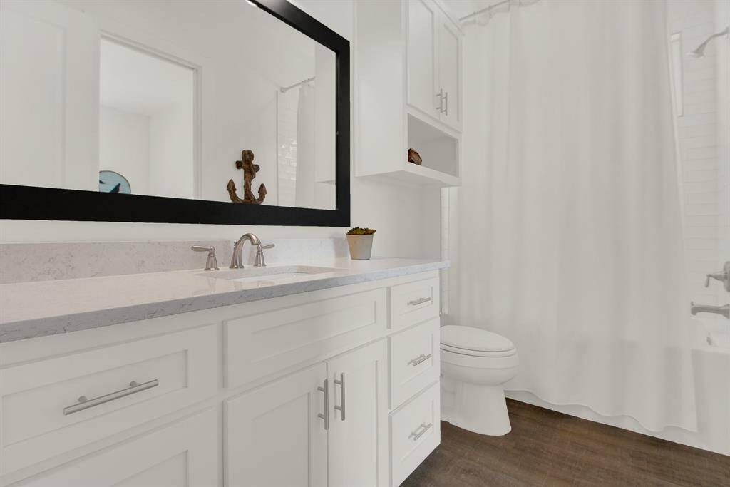 A modern, bright bathroom with a white color scheme, featuring a vanity with a granite countertop and undermount sink, a large mirror, a toilet, and a shower with a curtain. There's also built-in shelving for storage.