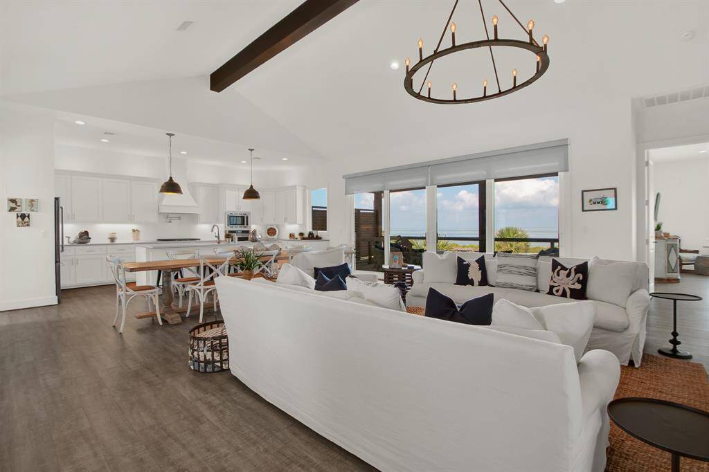 When you ascend the stairs a bright, open-plan living space featuring a modern kitchen with white cabinetry and integrated appliances, a dining area, and a comfortable living room welcomes you in. The room boasts high ceilings with exposed beams, elegant lighting fixtures, and large windows offering a view of the outdoors.
