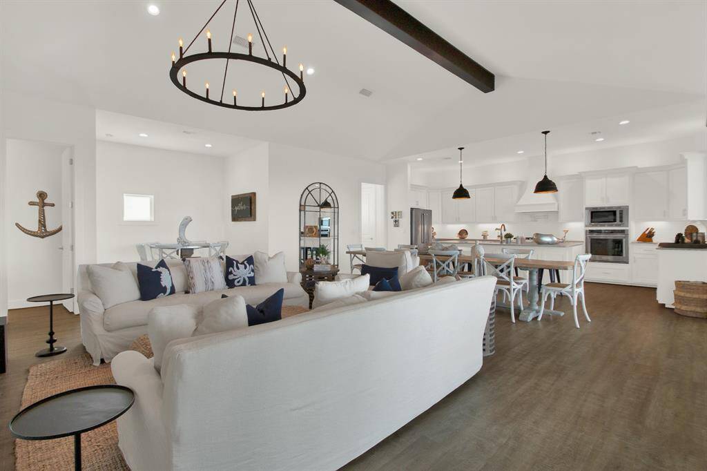 This open-concept living space features a spacious living area offering enough space for multiple pieces of furniture for a large amount of guests or a smaller intimate gathering. The room is well-lit by natural light and an elegant chandelier, complemented by dark wood beams and luxury vinyl plank floors.