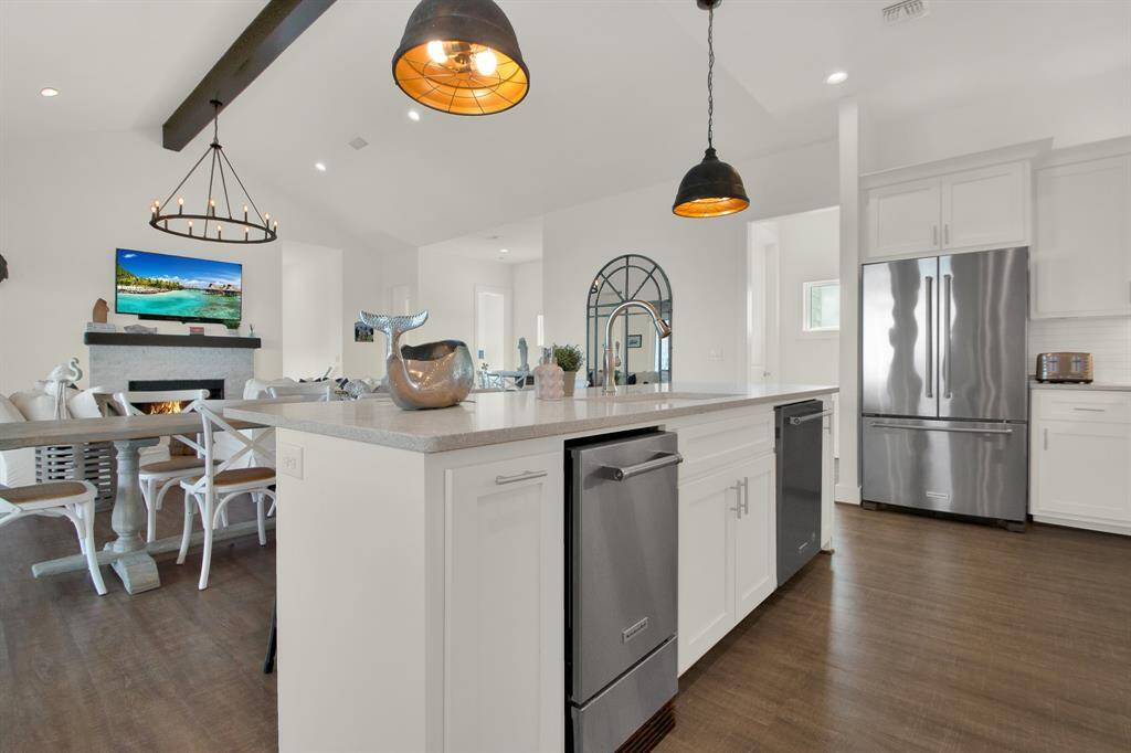 This is a bright and modern open-concept kitchen with a large center island, stainless steel appliances, and white cabinetry, leading into a cozy dining area with a fireplace and stylish lighting fixtures.