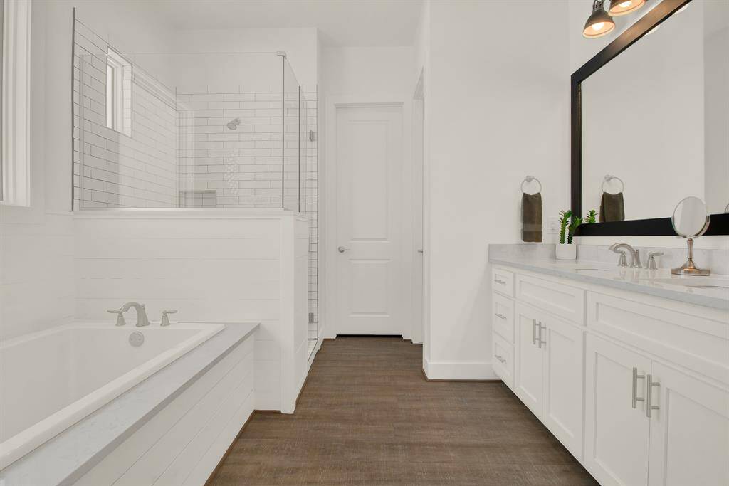 The primary bathroom is modern featuring a built-in bathtub, a glass-enclosed walk-in shower, a double vanity with ample storage, and a large mirror with overhead lighting. Clean lines, white cabinetry, and a neutral color palette give the space a bright and airy feel.