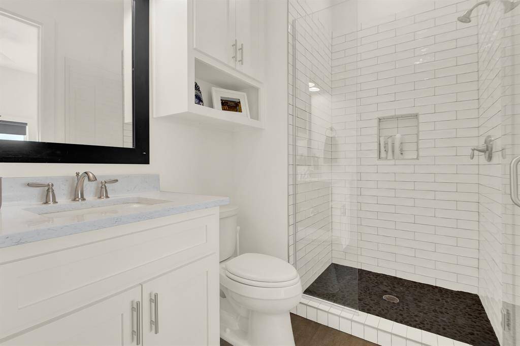 Modern bathroom featuring a white vanity with a granite countertop, under-mount sink, and a large mirror. There is a walk-in shower with white subway tiles and a pebble floor. The space is completed with a white toilet and built-in shelving for storage.