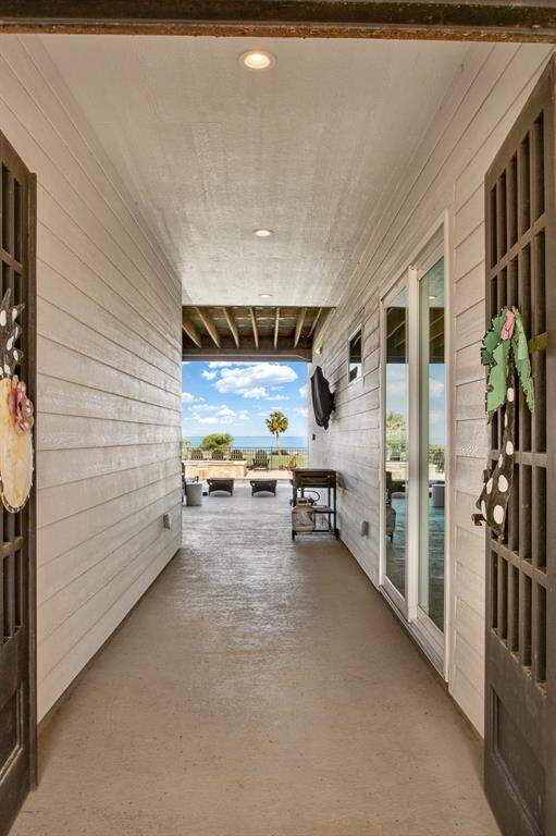 This is a covered, outdoor passageway with a modern aesthetic, featuring sleek lines, a concrete floor, and recessed lighting. Large sliding glass doors on one side provide entry into the first level of the home. The space leads to an open patio area with outdoor furniture and a clear view of the surroundings, which include palm trees and a blue sky, suggesting a pleasant, temperate climate.