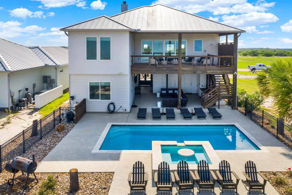 The back view shows the magnificent two-story home with two spacious covered areas, a private pool with a hot tub, and an outdoor entertainment area. The property offers a fenced backyard with ample space for relaxation and views of the surrounding landscape.