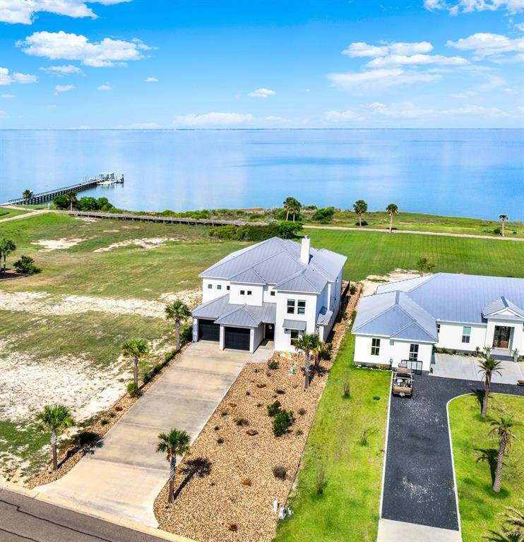 This is an aerial view of the front of the home with a gray metal roof and attached garage, situated in a spacious, grassy lot with direct access to a tranquil body of water. The property features a long public pier extending into the water and is neighbored by similar upscale homes, offering a serene and exclusive living experience.