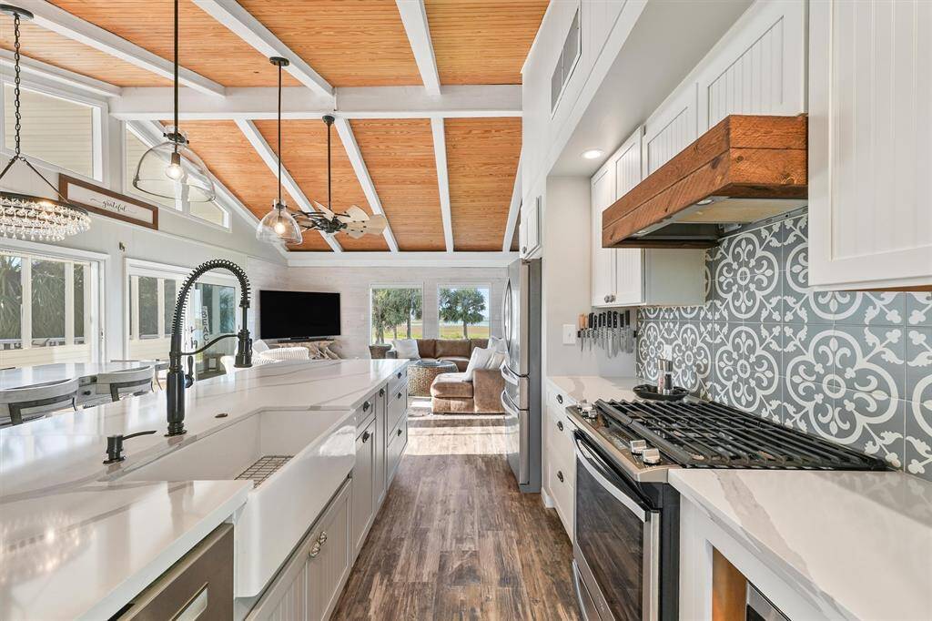 This bright, open-concept kitchen and living area features vaulted wooden ceilings, modern pendant lighting, and a stylish patterned backsplash. The space is complemented by a large island with a farmhouse sink, stainless steel appliances, and ample natural light from expansive windows, offering a cozy yet contemporary feel.