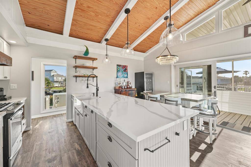 This spacious kitchen features a high, wood-paneled vaulted ceiling with exposed beams, a large central island with a sleek countertop, and modern pendant lighting. It opens to a dining area with ample natural light from large windows and a sliding glass door leading to an outdoor deck. The flooring is a warm-toned wood, and the decor is contemporary with a touch of elegance.