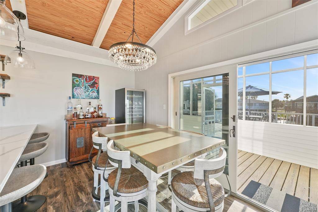 This bright dining area features a modern chandelier, a large central table with seating, and a stylish bar area. It opens to a deck through sliding glass doors, offering a view of the outdoors. The room has high ceilings with exposed beams and a warm wood finish.