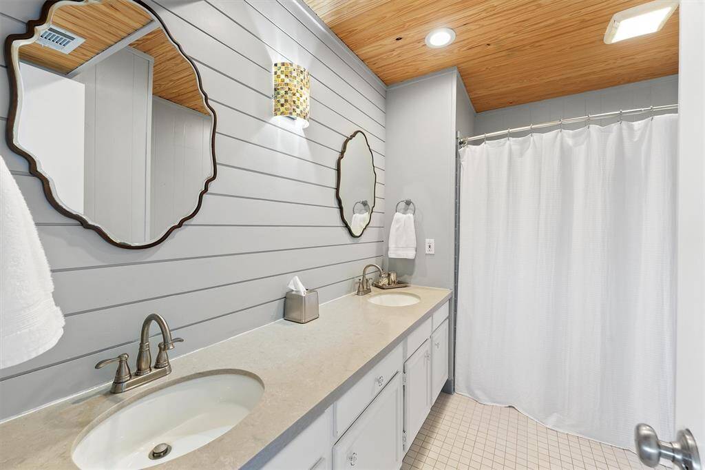 This  en-suite bathroom features a modern design with a double sink vanity, stylish mirrors, and a light wood-paneled ceiling. It includes a shower with a white curtain and soft, neutral tones for a fresh, inviting atmosphere.