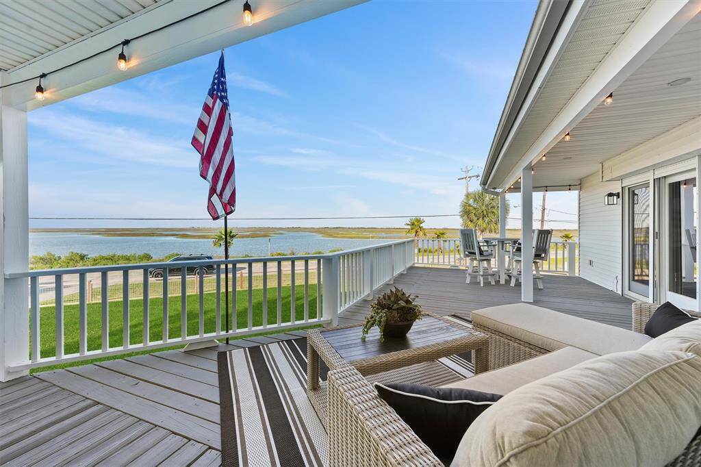 The photo showcases a spacious, covered deck with comfortable seating and dining area, offering stunning water views. The area is adorned with string lights and a USA flag, providing a cozy and inviting outdoor space perfect for relaxation and entertaining over looking the Bay!   No better sun rises anywhere.