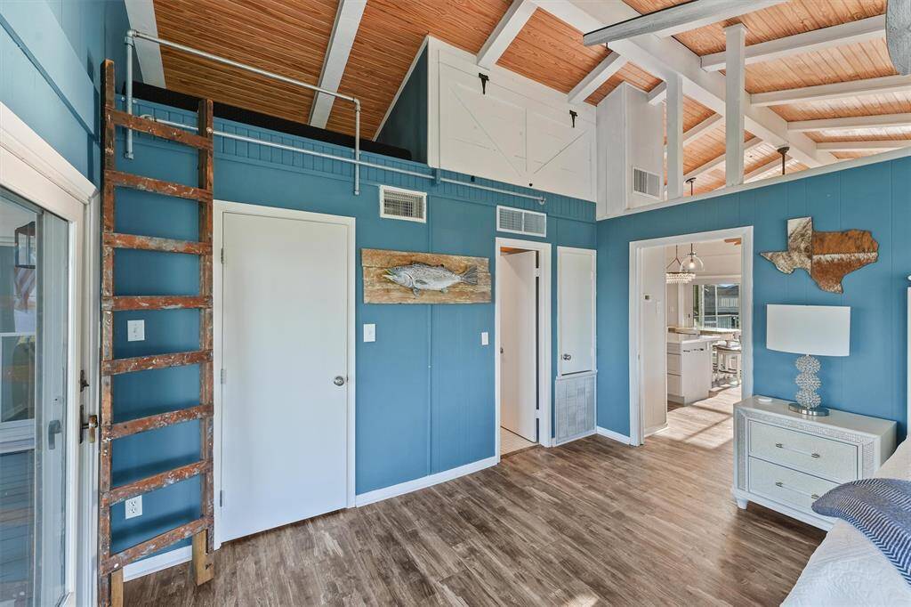 This 2nd primary features vaulted ceilings with exposed beams, wood flooring.. It includes a rustic wooden ladder leading to a loft space, a sliding glass door leading to the front veranda, and access to an open kitchen area. Decor includes coastal-themed artwork.