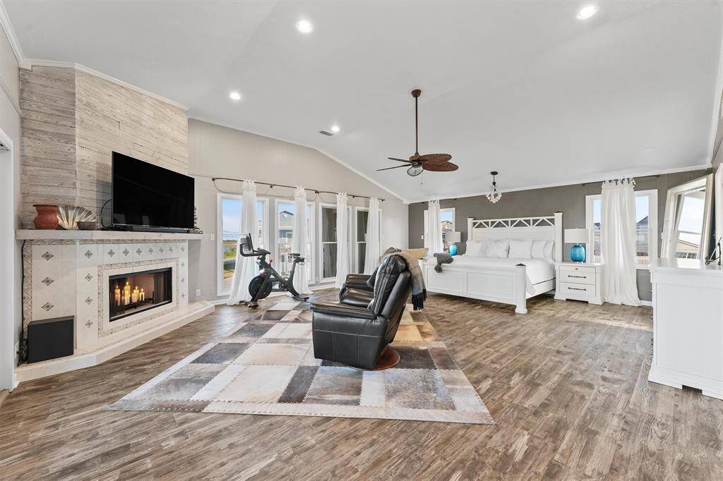 This spacious Primary bedroom features elegant wood flooring, a modern fireplace with a mounted TV, and ample natural light from large windows. It includes a cozy sitting area creating a luxurious and comfortable retreat.