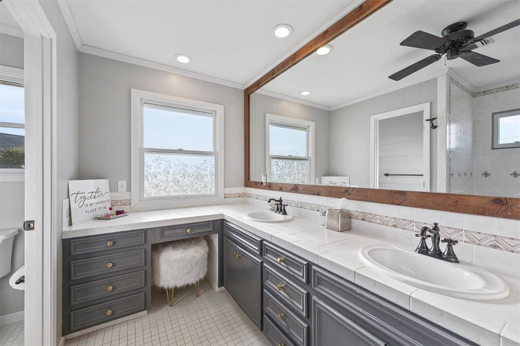This bathroom features a double sink vanity with stylish dark cabinetry and white countertops. It has a large mirror, a mounted ceiling fan, and ample natural light from two windows. The room also includes a decorative tiled backsplash and a cozy seating area.