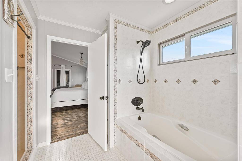 This photo shows a bright bathroom featuring a bathtub with a showerhead, accented by decorative tiles. A window provides natural light, and the space connects to a bedroom with wood flooring.