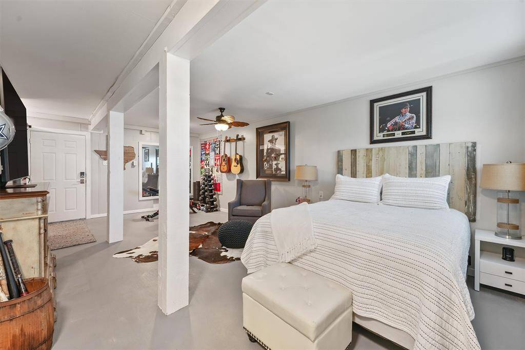 This spacious, well-lit room features a cozy bed with a rustic headboard, a ceiling fan, and stylish decor. It has a seating area with armchairs, musical instruments on display, and a modern aesthetic, making it a versatile living space.