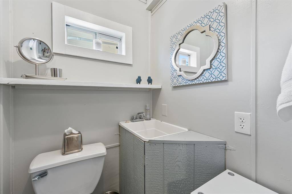 This is a compact bathroom with a modern design. It features a toilet and a small sink with a textured cabinet. The walls are light-colored, enhancing the natural light from a small window. A stylish mirror adds character, and a shelf offers space for toiletries.