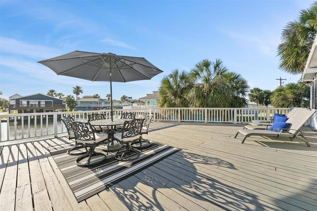 Spacious waterfront deck with elegant outdoor dining set under a large umbrella, two loungers, and scenic views of water and palm trees. Ideal for relaxing and entertaining.