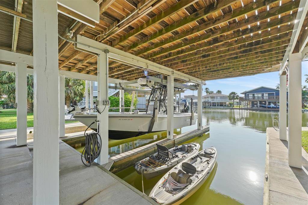 This photo showcases the covered boat dock with a lift, accommodating a large boat and two kayaks, set on a calm waterfront. The area is surrounded by lush greenery and offers a scenic view of neighboring waterfront homes. Ideal for boating enthusiasts.
