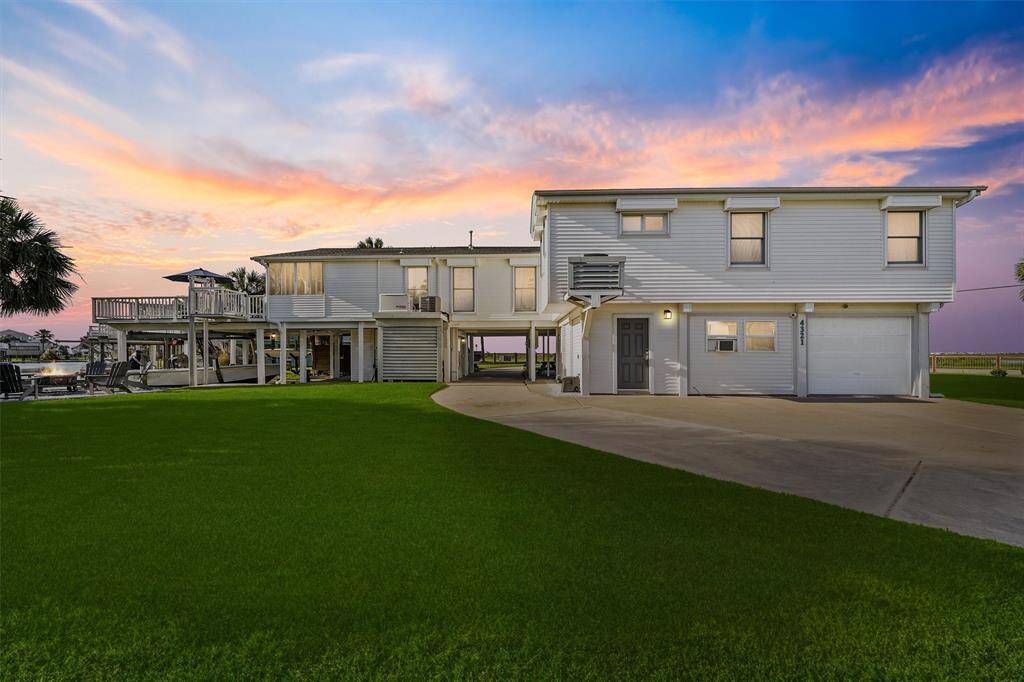 This photo showcases a spacious, elevated home with a modern exterior, featuring a large driveway and lush green lawn. The sunset sky adds a warm, inviting atmosphere, highlighting the property's ample outdoor space and potential for relaxation and entertainment.