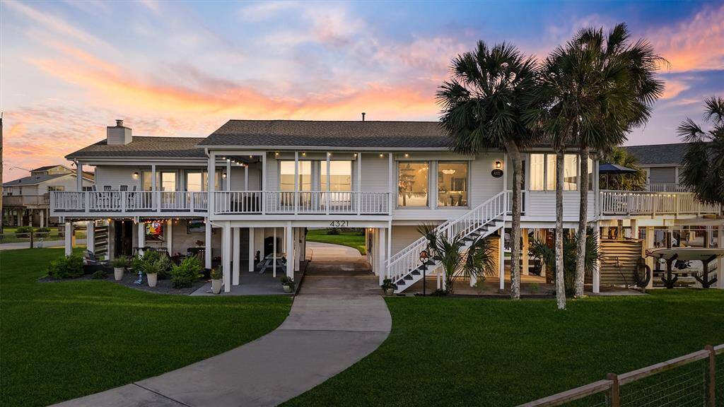 This elevated coastal-style home features a spacious wraparound deck, a lush green lawn, and striking palm trees. The sunset backdrop enhances its inviting ambiance, perfect for relaxation and outdoor enjoyment.