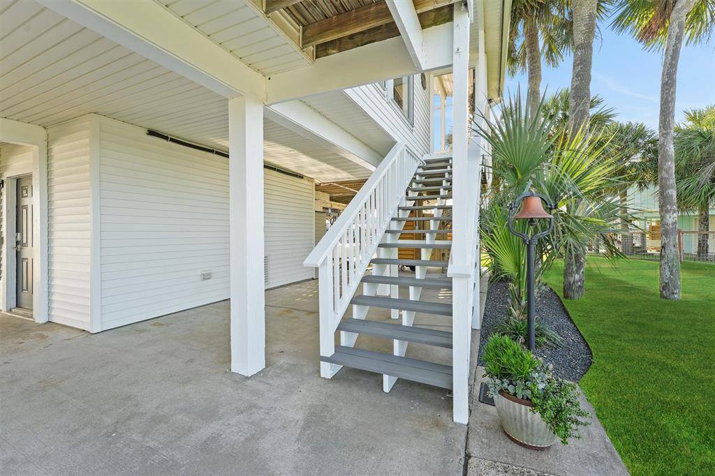 This photo showcases a raised home with a covered area beneath, featuring a staircase leading to the main entrance. The exterior is clad in white siding, and the surrounding landscape includes well-maintained grass and palm trees, offering a tropical feel.