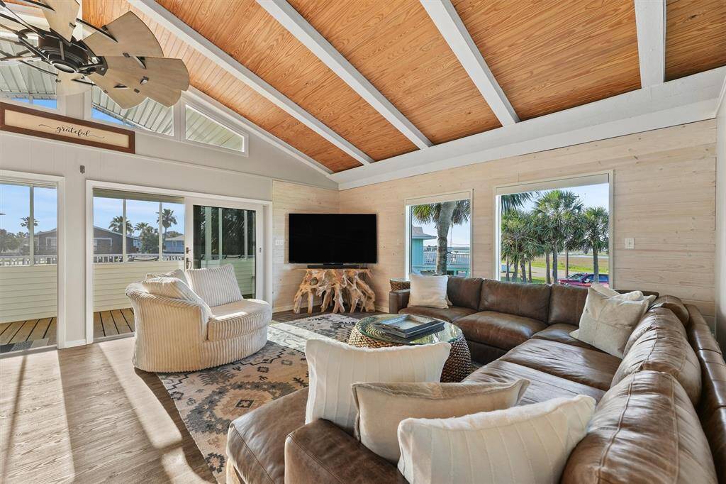 This inviting living room features high vaulted ceilings with wood accents and natural light streaming through multiple windows. It opens to a spacious deck, offering views of gorgeous canal sunsets, creating a warm and cozy atmosphere.