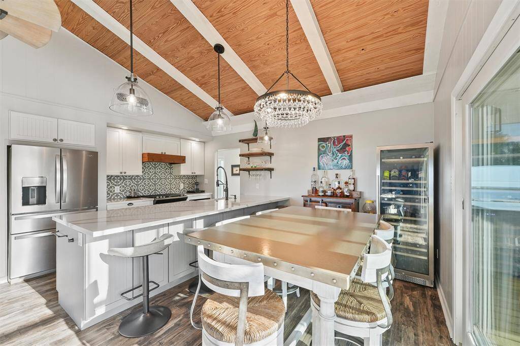 This kitchen features a modern, open design with a high, wood-beamed ceiling and stylish pendant lighting. It includes a large island with bar seating, stainless steel appliances, and a decorative tile backsplash. A bar area add a touch of luxury, while the sliding glass doors to the large balcony  offers plenty of natural light.