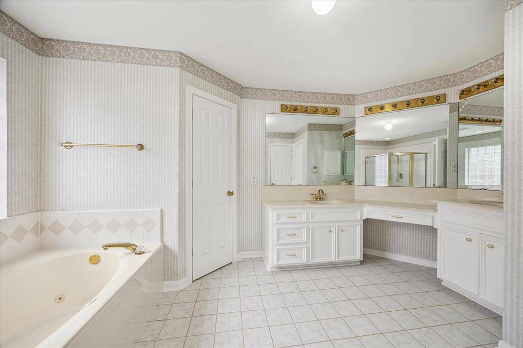 The primary bathroom features a spacious layout with a large corner vanity, ample counter space, and multiple mirrors. It includes a luxurious bathtub, light tile flooring, and tasteful wallpaper, creating a bright and inviting atmosphere.
