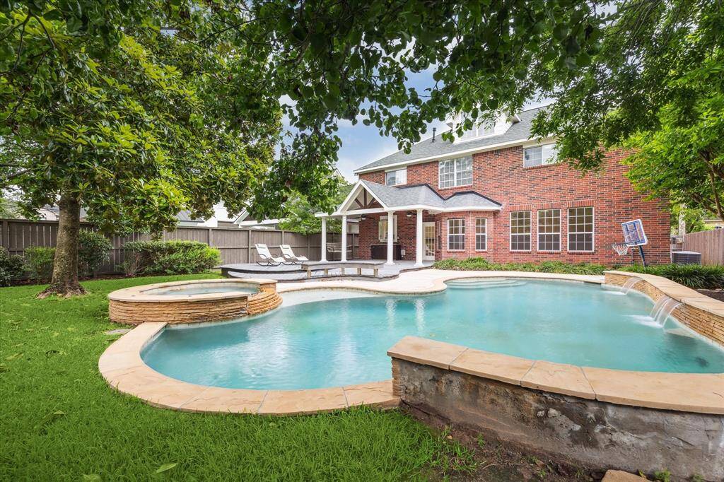 This beautiful backyard features a spacious pool with a waterfall, surrounded by lush greenery. There is a cozy covered patio area, perfect for relaxation and outdoor entertaining.