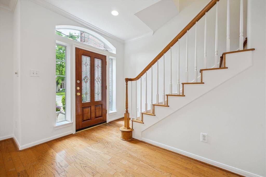 This inviting entryway features a wooden door with decorative glass panels, flanked by tall windows for natural light. The warm hardwood floor complements the wooden staircase with white balusters, creating a welcoming atmosphere.