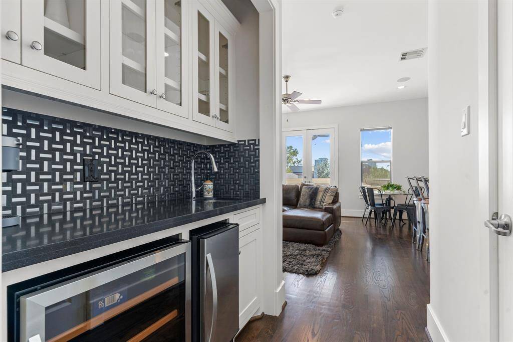 Enjoy the convenience of the game room's wet bar, complete with white glass-front cabinets, a stone countertop, stainless sink, wine refrigerator, and a gray subway tile backsplash for an elegant touch.